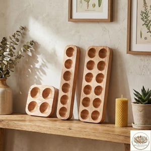 May include: Wooden egg holders in various sizes. The holders are made of light-colored wood and feature circular indentations to hold eggs. One holder is square with four holes, while two are rectangular with space for 10 and 12 eggs. A candle and a potted plant are also visible.