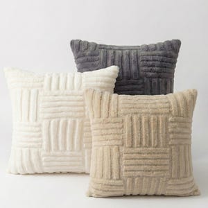 Puede incluir: Tres cojines decorativos cuadrados en crema, gris y beige. Cada cojín tiene un patrón texturizado y tejido. Los cojines están dispuestos en grupo, con el cojín crema delante, el cojín gris detrás y el cojín beige a la derecha.