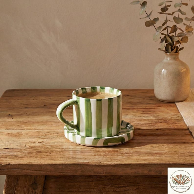 May include: A ceramic coffee cup and saucer set with vertical green stripes on a white background. The cup is filled with coffee. The set is on a wooden table, with a small vase of greenery in the background.