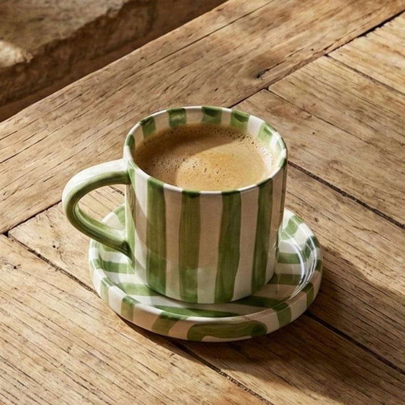 May include: A ceramic coffee cup and saucer set with a green and white striped pattern. The cup is filled with coffee and sits on a wooden table. The handle of the cup is also green and white striped.
