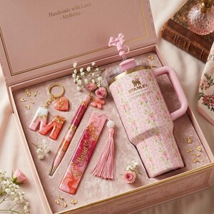 May include: A pink gift box with a floral-patterned Stanley Loveshack Fancy tumbler, pen, bookmark, keychain, and tassel. The tumbler has a handle and straw. The box includes the text "Handmade with Love - MyBetsy -" and is decorated with gold accents and small flowers.