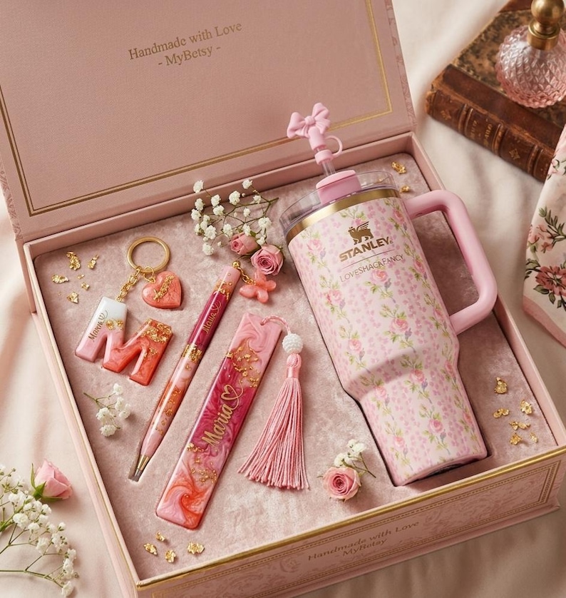 May include: A pink gift box with a floral-patterned Stanley Loveshack Fancy tumbler, pen, bookmark, keychain, and tassel. The tumbler has a handle and straw. The box includes the text "Handmade with Love - MyBetsy -" and is decorated with gold accents and small flowers.