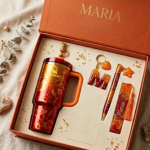 以下が含まれることがあります： MARIAと書かれたオレンジ色の箱に入ったギフトセット。パイナップルのトッパーが付いた赤と金のフローラル柄のスタンレー製タンブラー、ハートのチャームが付いたMのキーホルダー、蝶のチャームが付いたペン、オレンジ色のタッセルが付いたしおりが含まれています。