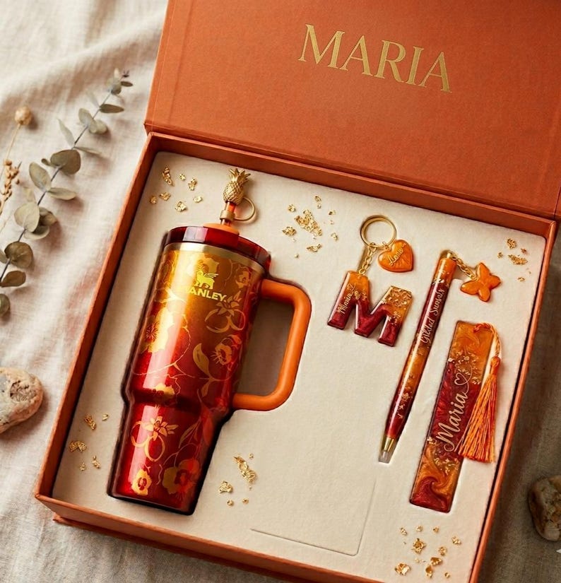 以下が含まれることがあります： MARIAと書かれたオレンジ色の箱に入ったギフトセット。パイナップルのトッパーが付いた赤と金のフローラル柄のスタンレー製タンブラー、ハートのチャームが付いたMのキーホルダー、蝶のチャームが付いたペン、オレンジ色のタッセルが付いたしおりが含まれています。