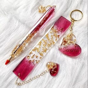 May include: A set of handmade resin accessories, including a pen, bookmark, keychain, and charm. The items are a gradient of red and clear resin, embedded with gold flakes. The pen and keychain have the name "Gemma" written on them.