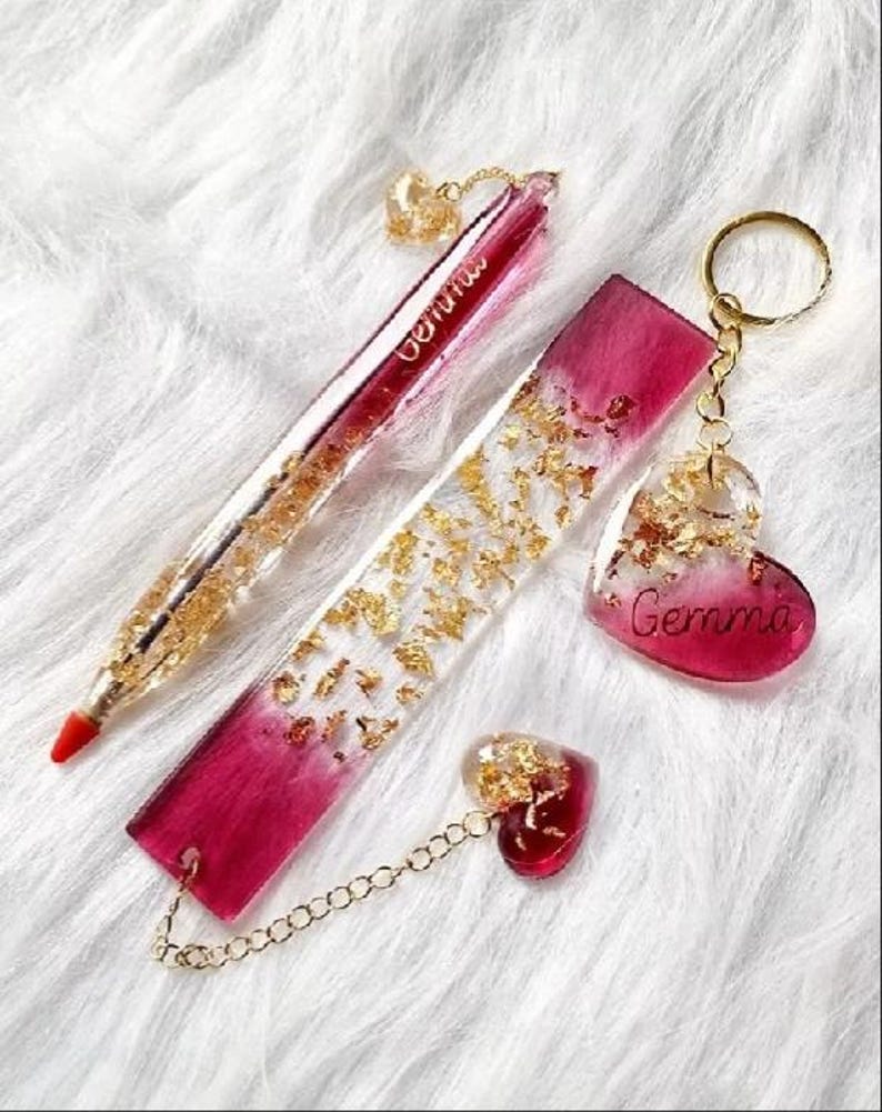 May include: A set of handmade resin accessories, including a pen, bookmark, keychain, and charm. The items are a gradient of red and clear resin, embedded with gold flakes. The pen and keychain have the name "Gemma" written on them.