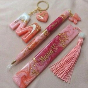 May include: A collection of pink and gold resin accessories, including a pen, bookmark, keychain, and letter "M". The pen and bookmark are inscribed with the name "Maria". The items feature gold flakes and a tassel.