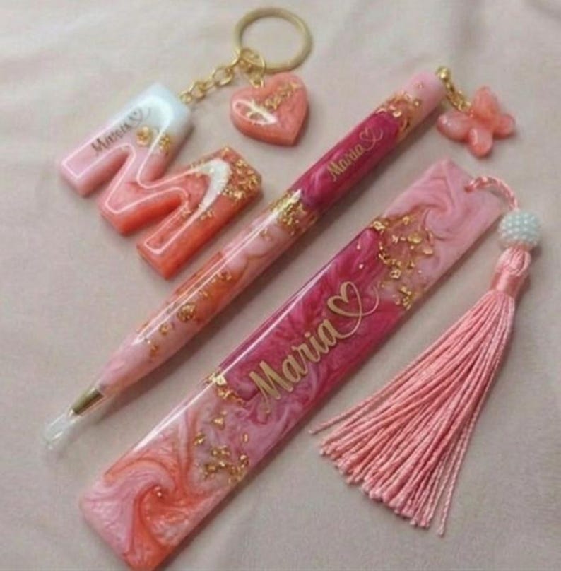 May include: A collection of pink and gold resin accessories, including a pen, bookmark, keychain, and letter "M". The pen and bookmark are inscribed with the name "Maria". The items feature gold flakes and a tassel.