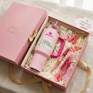 May include: A pink gift set in a pink box includes a Stanley Quencher H2.0 tumbler, a pen, a bookmark, and keychains. The box is tied with a gold ribbon and contains the text "Maria's Curated Creations". The gift set is designed for Maria.