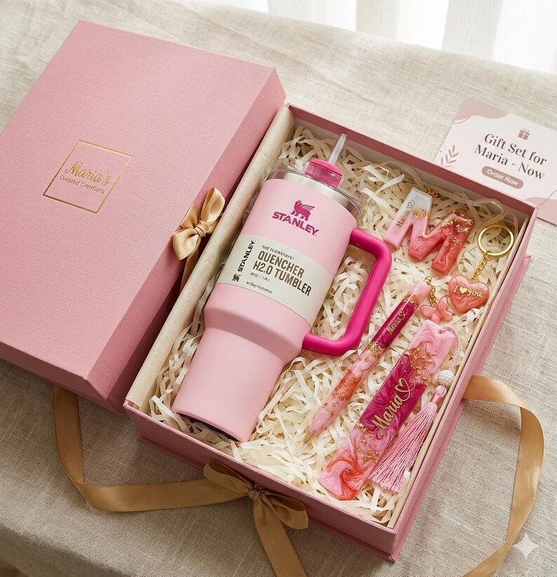 May include: A pink gift set in a pink box includes a Stanley Quencher H2.0 tumbler, a pen, a bookmark, and keychains. The box is tied with a gold ribbon and contains the text "Maria's Curated Creations". The gift set is designed for Maria.