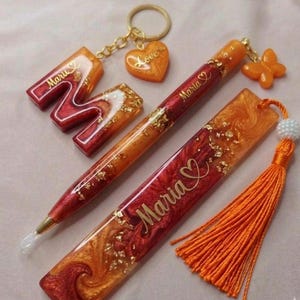 以下が含まれることがあります： 名前Maruが入ったM字型のキーホルダー、名前Mariaが入ったペン、名前Mariaが入った長方形のしおりのレジンセット。各アイテムは、赤とオレンジのマーブル模様の仕上げで、金箔と金色の文字が施されています。