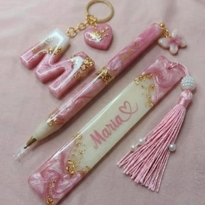May include: A collection of pink and gold resin accessories, including a letter "M" keychain, a heart-shaped keychain, a pen, and a bookmark with the name "Maria" and a heart symbol. The items have a marbled effect and gold flakes.