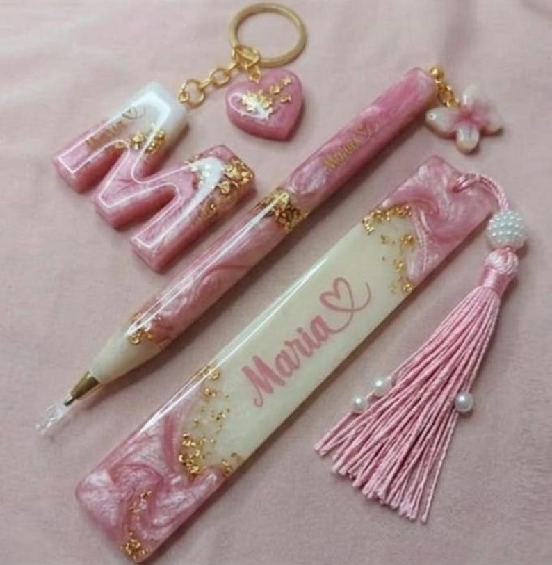 May include: A collection of pink and gold resin accessories, including a letter "M" keychain, a heart-shaped keychain, a pen, and a bookmark with the name "Maria" and a heart symbol. The items have a marbled effect and gold flakes.