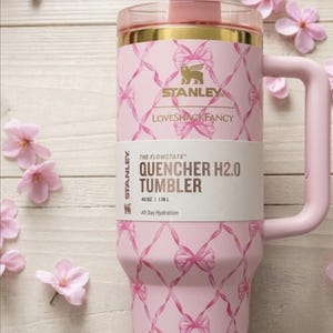 Op de afbeelding: Een lichtroze Stanley Quencher H2.0 beker met een inhoud van 1,18 L, voorzien van een herhalend patroon van roze linten. De beker heeft een goudkleurige rand, een bijpassend roze handvat, een doorzichtig rietje en een label met de tekst.