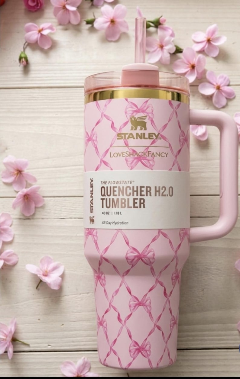 Op de afbeelding: Een lichtroze Stanley Quencher H2.0 beker met een inhoud van 1,18 L, voorzien van een herhalend patroon van roze linten. De beker heeft een goudkleurige rand, een bijpassend roze handvat, een doorzichtig rietje en een label met de tekst.