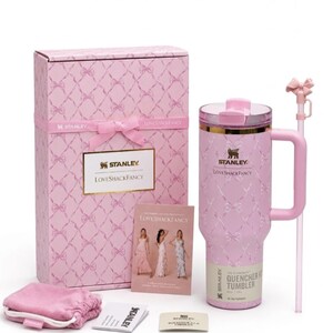 Op de afbeelding: Een lichtroze Stanley Quencher beker met een wit strikpatroon, een bijpassende roze doos met strik, een rietje met strikopzetstuk, een klein roze tasje en een LoveShackFancy informatiekaart. De beker is voorzien van een gouden rand en handvat.