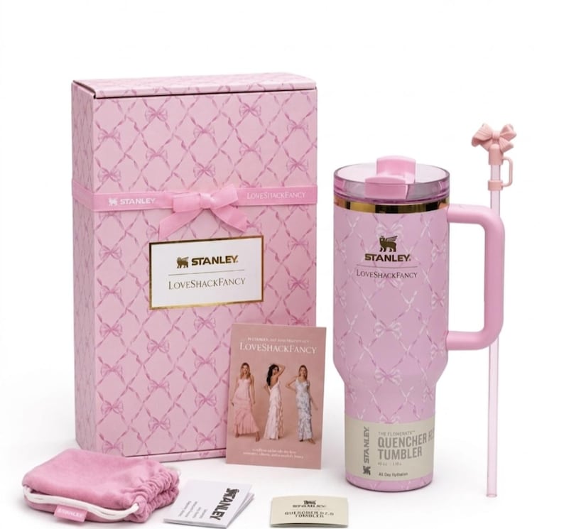 Op de afbeelding: Een lichtroze Stanley Quencher beker met een wit strikpatroon, een bijpassende roze doos met strik, een rietje met strikopzetstuk, een klein roze tasje en een LoveShackFancy informatiekaart. De beker is voorzien van een gouden rand en handvat.