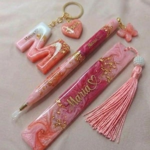 May include: A collection of pink and gold resin accessories, including a keychain with a heart charm, a pen, a bookmark with a tassel, and a letter "M". The items feature gold flakes and the name "Maria".