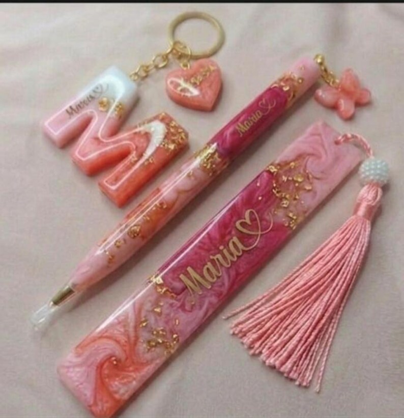 May include: A collection of pink and gold resin accessories, including a keychain with a heart charm, a pen, a bookmark with a tassel, and a letter "M". The items feature gold flakes and the name "Maria".