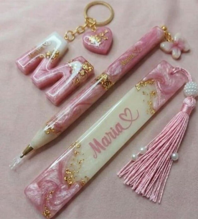 May include: A collection of pink resin accessories, including a pen, bookmark, keychain, and letter "M". The items feature gold leaf accents and the name "Maria". The bookmark has a pink tassel and pearl beads.