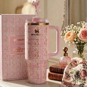 May include: A pink floral-patterned Stanley x Loveshackfancy 40oz Quencher H2.0 tumbler with a handle and a bow-shaped straw topper. The tumbler is next to a matching box. The background includes flowers and a decorative perfume bottle.