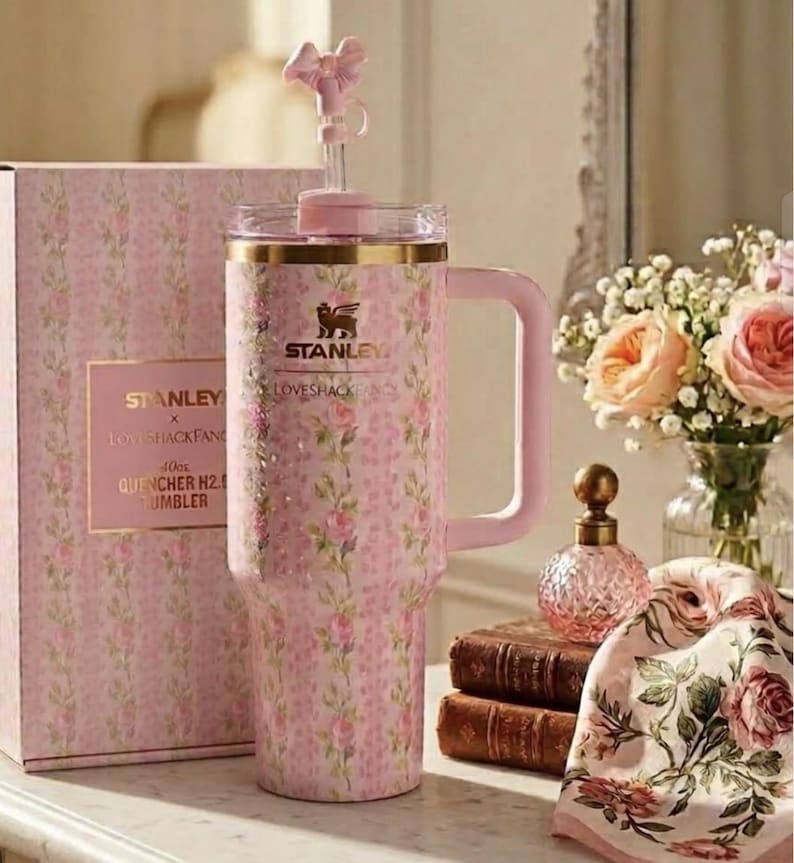 May include: A pink floral-patterned Stanley x Loveshackfancy 40oz Quencher H2.0 tumbler with a handle and a bow-shaped straw topper. The tumbler is next to a matching box. The background includes flowers and a decorative perfume bottle.