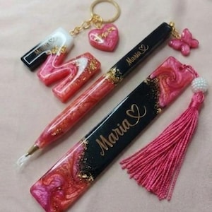 May include: A collection of handmade resin accessories in shades of pink, red, and gold. Items include a pen, bookmark, keychain with a heart charm, and a letter "M" with the name "Maria" on some items. A pink tassel is also included.