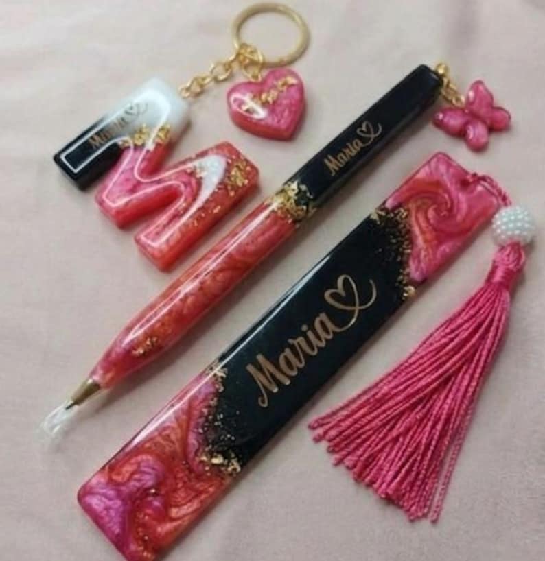 May include: A collection of handmade resin accessories in shades of pink, red, and gold. Items include a pen, bookmark, keychain with a heart charm, and a letter "M" with the name "Maria" on some items. A pink tassel is also included.
