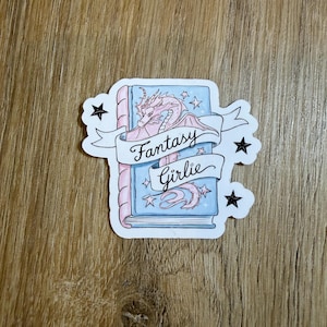May include: A sticker featuring a fantasy-themed design. The sticker depicts a book with a pink dragon and the words "Fantasy Girlie" on a banner. The book is blue with pink accents and surrounded by stars and black star shapes.