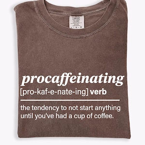 Puede incluir: Una camiseta marrón doblada con la palabra "procaffeinating" en blanco, su pronunciación y definición. La definición dice: "la tendencia a no empezar nada hasta que hayas tomado una taza de café."