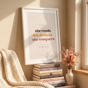 Peut inclure: Une affiche encadrée avec le texte "she reads, she dreams, she conquers" en marron, jaune et rose. L'affiche est posée sur une pile de livres, avec un vase de fleurs et une couverture douillette à proximité. Les livres sont empilés les uns sur les autres.