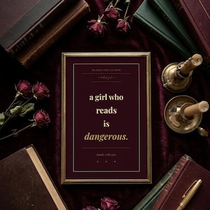 Peut inclure: Un cadre rectangulaire doré contenant une impression bordeaux foncé avec le texte doré : READER COLLECTION, a girl who reads is dangerous., handle with care. Le cadre est entouré de plusieurs livres anciens, de roses rouges séchées et d'un stylo doré.