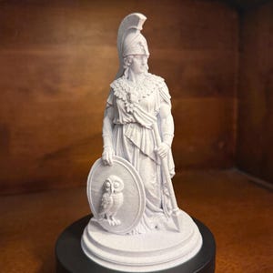 May include: A white matte statue of a figure wearing a crested helmet and draped clothing, holding a long spear in one hand and a round shield featuring a relief of an owl in the other, standing on a circular base atop a black cylindrical stand.