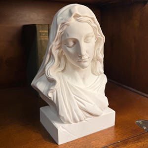 May include: A white matte bust sculpture depicting a figure with closed eyes and a draped head covering. The sculpture features flowing hair, a serene facial expression, and a draped garment over the shoulders, all set on a rectangular base.