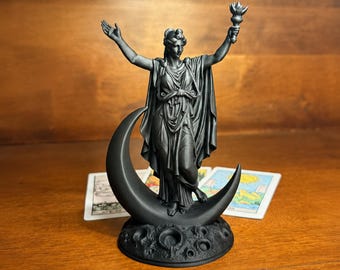 May include: A matte black figurine of a robed figure standing on a crescent moon base. The figure has one arm raised and the other holding a torch. The base features a textured, cratered surface. Two rectangular cards are visible behind the statue.