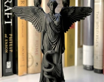 May include: A matte black figurine of a winged figure standing on a circular base. The figure wears draped robes and has an owl perched on each shoulder. A snake is coiled around the lower portion of the figure's body, extending from the waist down.