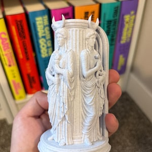 May include: A light gray, speckled 3D-printed figurine featuring two standing figures in draped robes, each wearing a crescent moon headpiece. One figure holds a torch, while the other holds a snake. They are positioned against a central pillar.