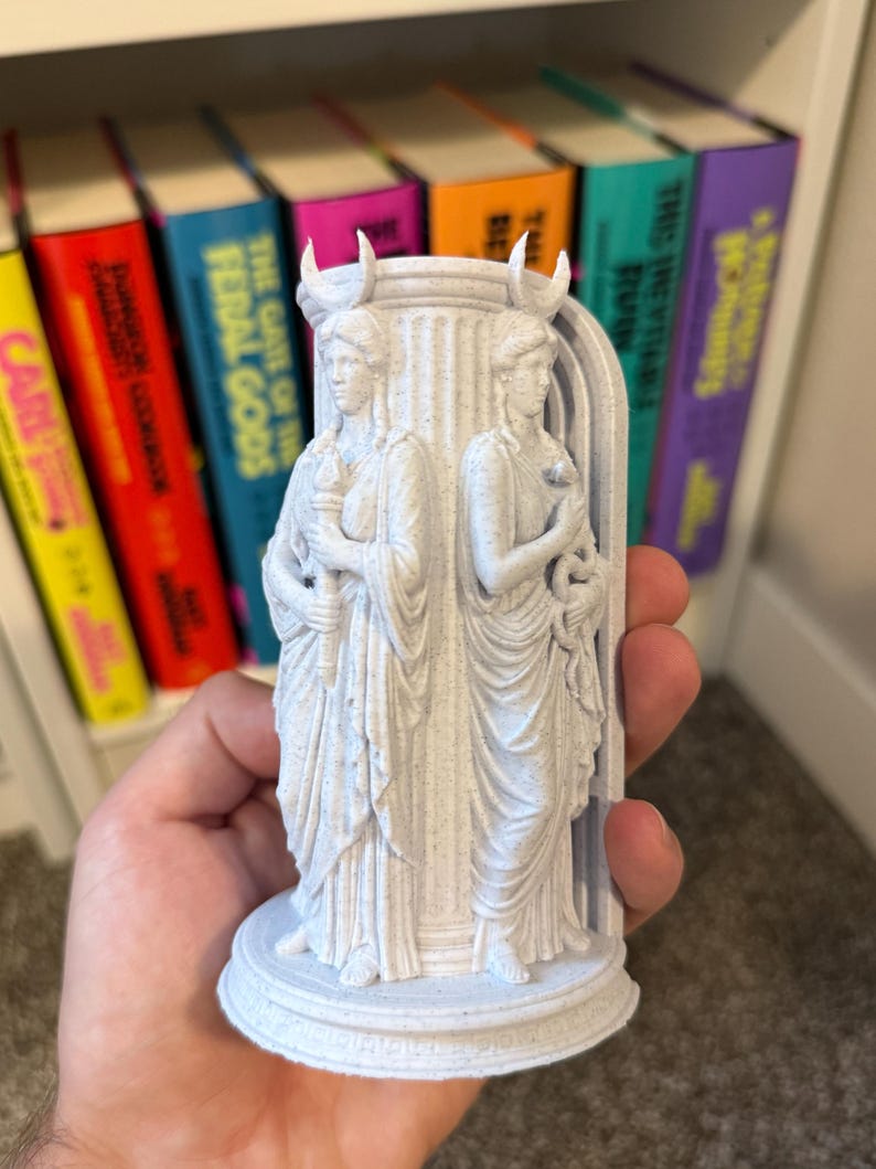 May include: A light gray, speckled 3D-printed figurine featuring two standing figures in draped robes, each wearing a crescent moon headpiece. One figure holds a torch, while the other holds a snake. They are positioned against a central pillar.