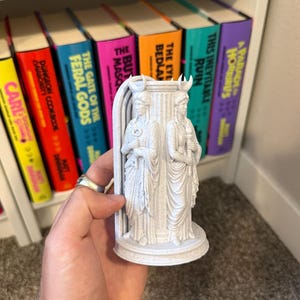 May include: A light gray 3D printed figurine featuring two standing figures with crescent moon headpieces, positioned on either side of a central pillar. One figure holds a key, while the other holds a torch, all set upon a circular base.