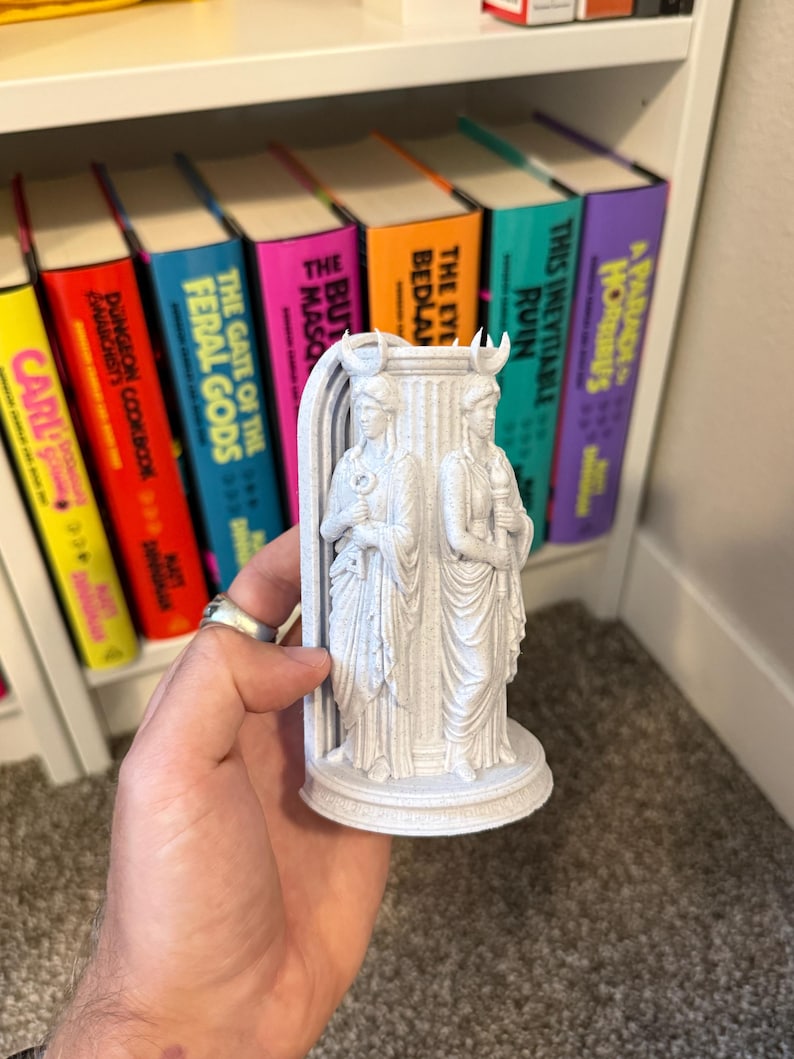 May include: A light gray 3D printed figurine featuring two standing figures with crescent moon headpieces, positioned on either side of a central pillar. One figure holds a key, while the other holds a torch, all set upon a circular base.