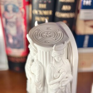 May include: A white, three-dimensional printed sculpture featuring a central fluted column base topped with a circular labyrinth design. Three identical figures with crescent moon headpieces are positioned around the column, each holding a small snake.