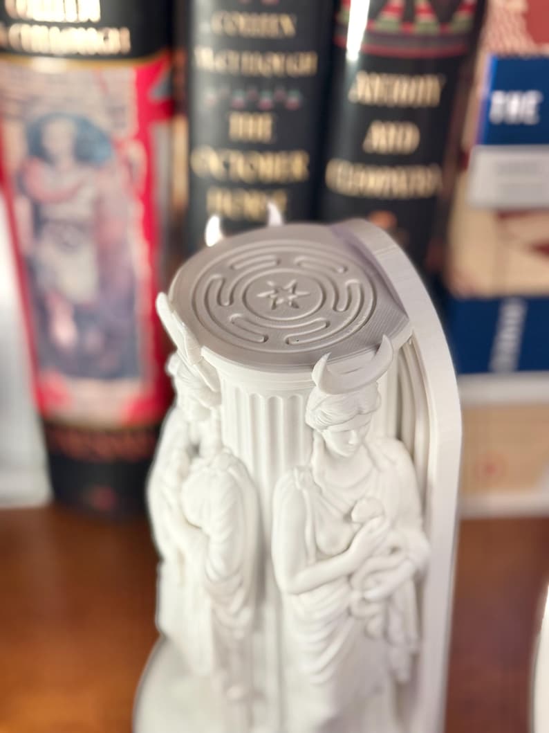 May include: A white, three-dimensional printed sculpture featuring a central fluted column base topped with a circular labyrinth design. Three identical figures with crescent moon headpieces are positioned around the column, each holding a small snake.
