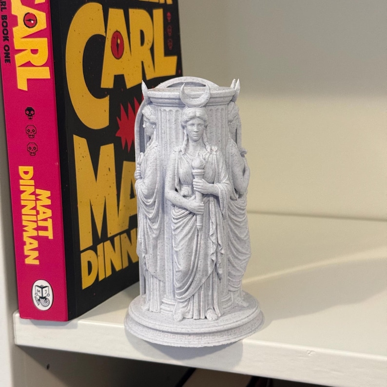May include: A light gray, 3D-printed cylindrical sculpture featuring three identical figures standing around a central pillar. Each figure wears a draped robe, holds a torch, and has a crescent moon emblem on their head. The base has a Greek key pattern.