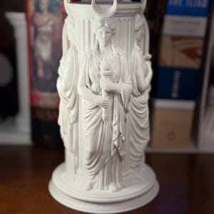 May include: A white cylindrical sculpture featuring three figures standing around a central column. Each figure wears draped robes and a crescent moon headpiece. One figure holds a torch. The base is circular with a tiered edge design.