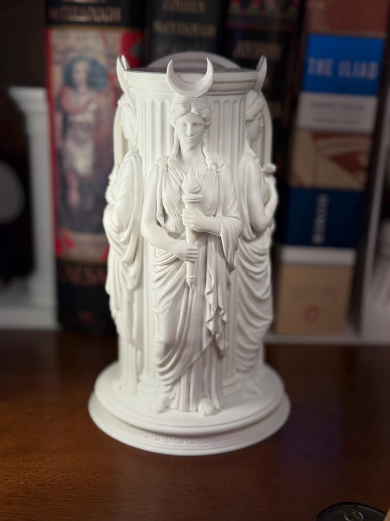 May include: A white cylindrical sculpture featuring three figures standing around a central column. Each figure wears draped robes and a crescent moon headpiece. One figure holds a torch. The base is circular with a tiered edge design.