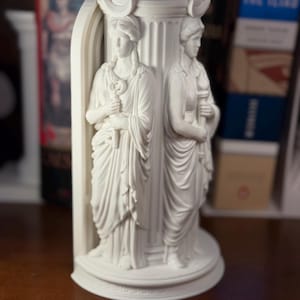 May include: A white matte bookend featuring two sculpted figures standing against a fluted column. Each figure wears a crescent moon headpiece and draped robes. One figure holds a key, while the other holds a torch, all set on a circular base.