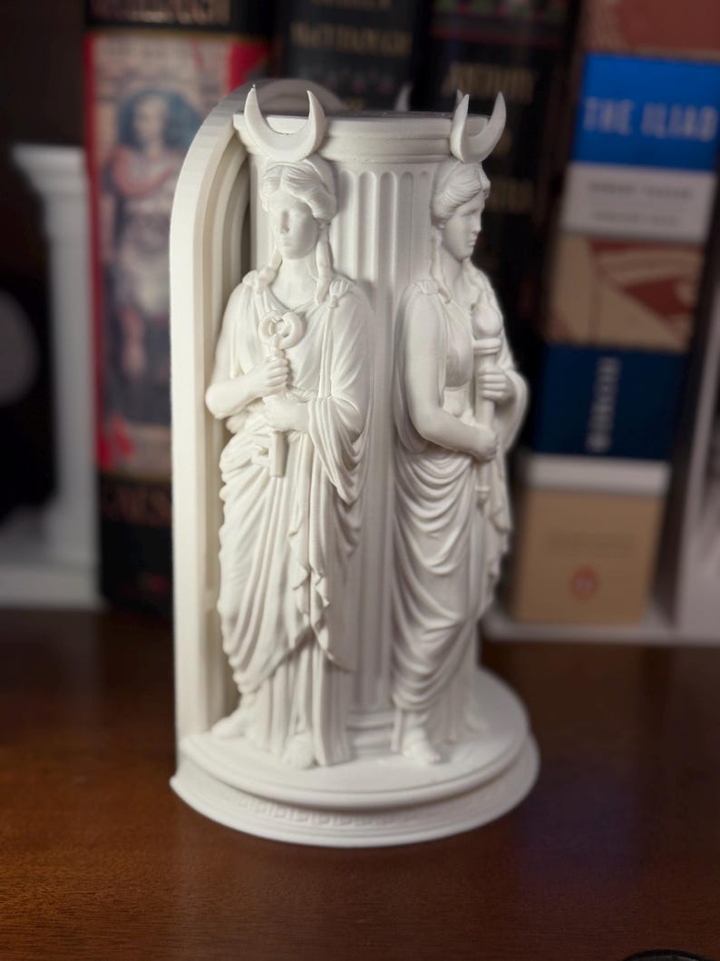 May include: A white matte bookend featuring two sculpted figures standing against a fluted column. Each figure wears a crescent moon headpiece and draped robes. One figure holds a key, while the other holds a torch, all set on a circular base.