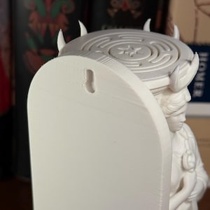 May include: A white 3D-printed wall-mounted sculpture featuring a figure with a classical hairstyle supporting a circular platform. The platform displays a labyrinth pattern with a central star. A keyhole slot is visible on the flat back panel.