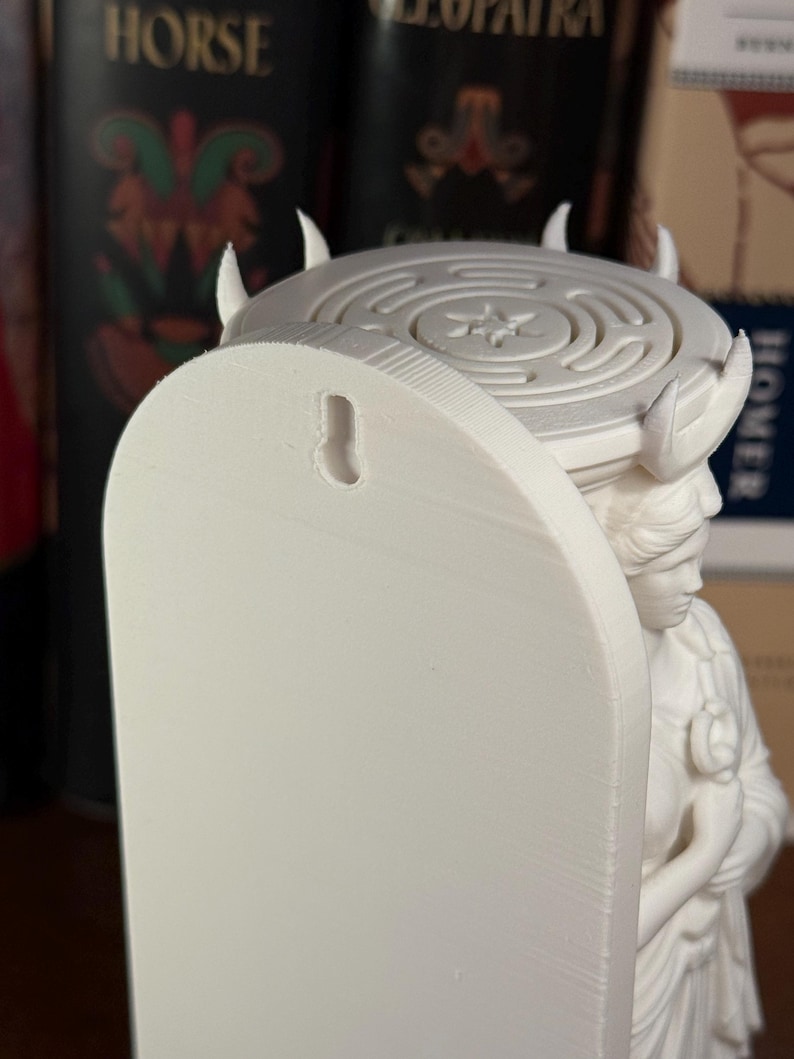 May include: A white 3D-printed wall-mounted sculpture featuring a figure with a classical hairstyle supporting a circular platform. The platform displays a labyrinth pattern with a central star. A keyhole slot is visible on the flat back panel.