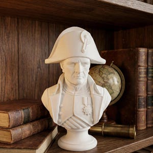 Może przedstawiać: Białe popiersie postaci historycznej, w dwurożnym kapeluszu i stroju wojskowym, stoi na półce. Otaczają je antyczne książki, globus i mosiężny teleskop, tworząc klasyczną, naukową estetykę.